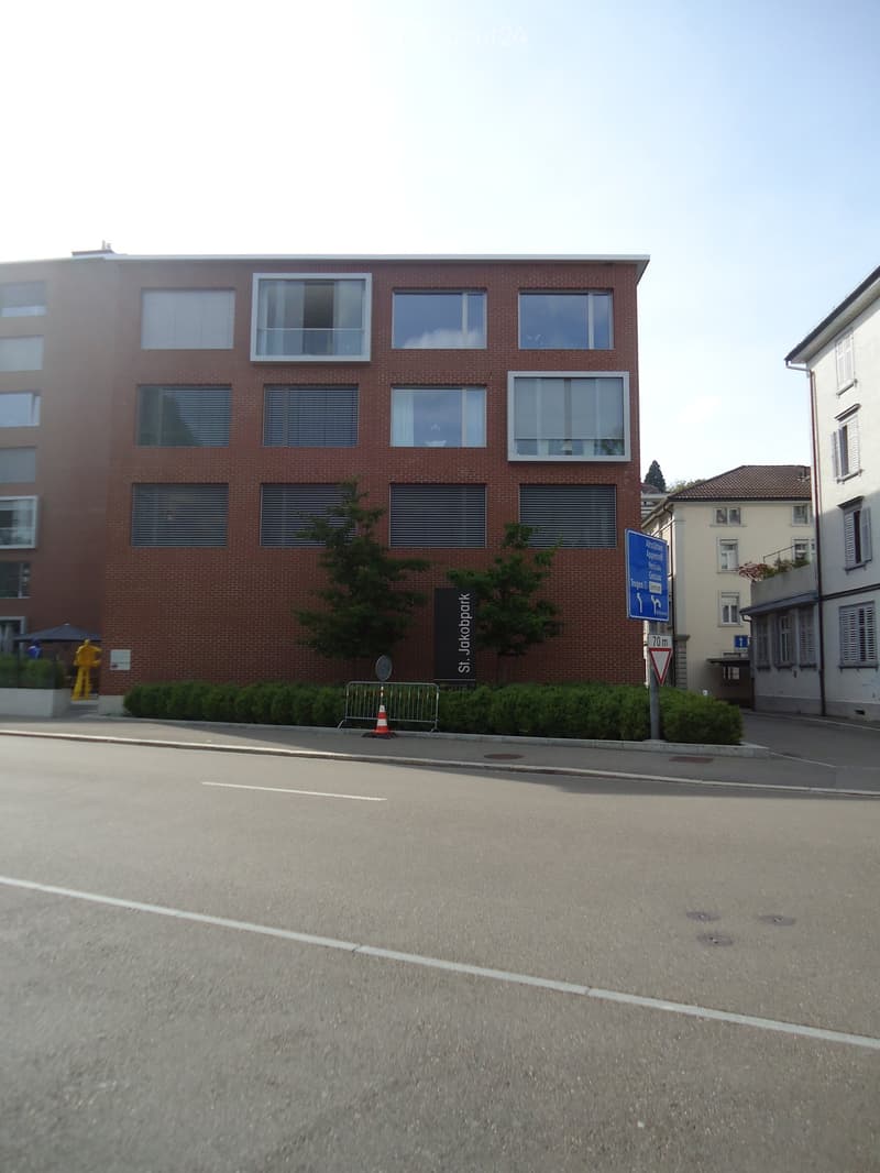 A multi-story brick building with large windows and a sign reading "St. Jakobpark" on the facade. The building is surrounded by greenery and a paved street.