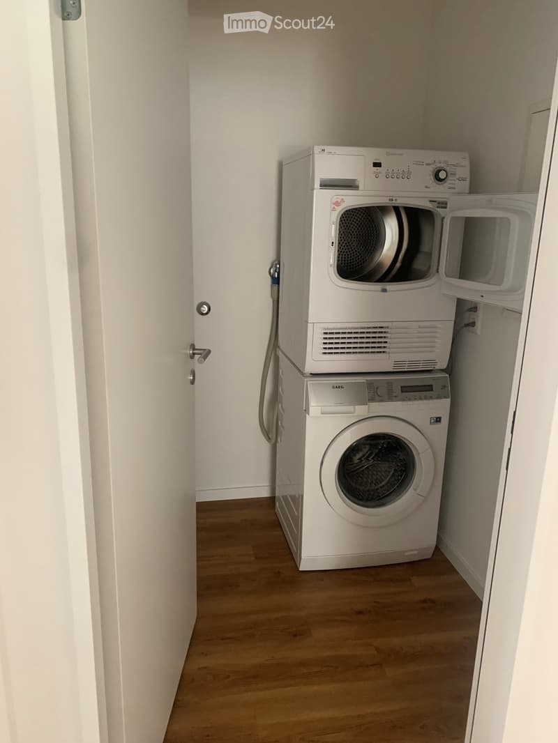 A small, well-lit room with wooden flooring and white walls, featuring a stacked washing machine and dryer unit.