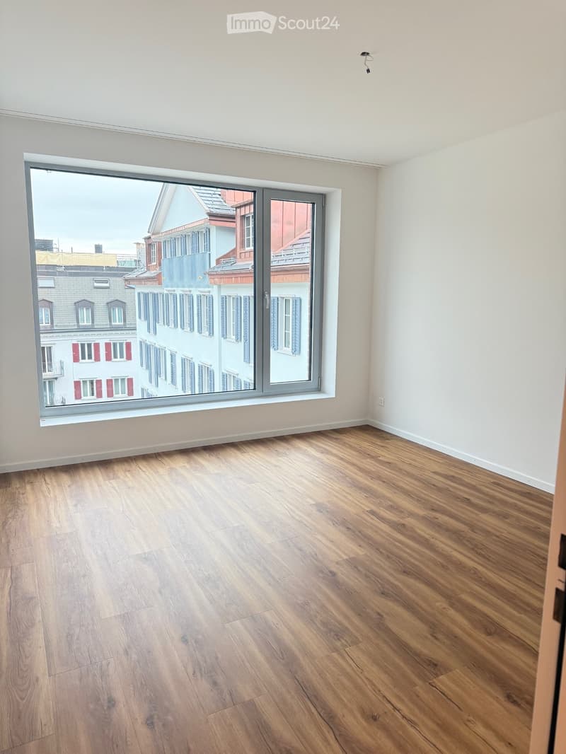 Empty room with wooden flooring and large window offering a view of neighboring buildings.