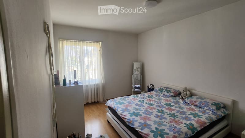 A well-lit bedroom with a wooden floor, a large window with white curtains, a bed with a colorful floral duvet, a mirror, a nightstand, and a small stool.