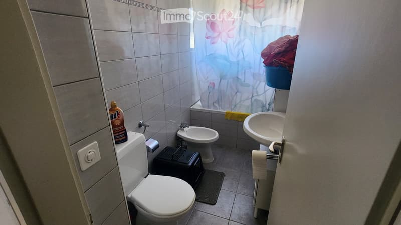 A compact bathroom with a toilet, sink, bidet, and shower area. The shower curtain features a floral design, and a bottle of cleaning solution is placed on the toilet tank. A small stool and a towel are also visible.