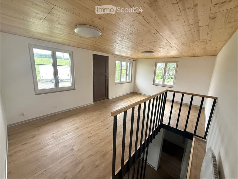 Empty room with wooden flooring, white walls, and a staircase leading downwards. Features include a large window, a door, and recessed lighting.