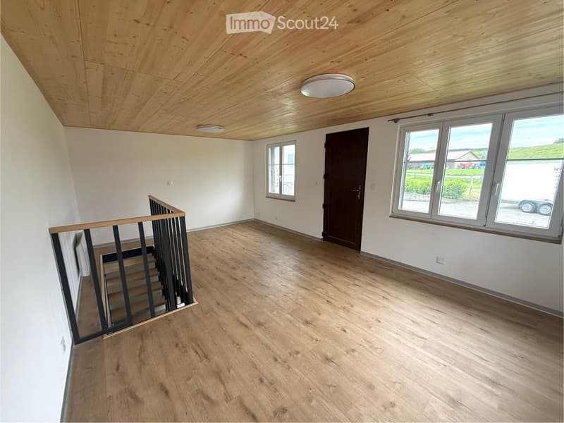 Empty room with wooden flooring, white walls, and a staircase leading upwards. Features large windows and a wooden ceiling.
