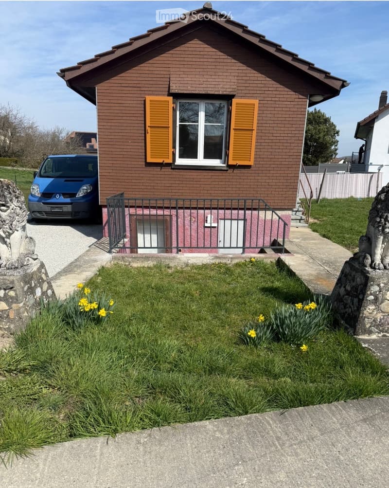 Two-story house with brown brick exterior, white-framed windows with orange shutters, a small garden with yellow flowers, and a blue van parked in the driveway.