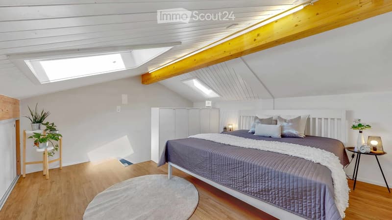 Bright, modern bedroom with wooden floors, white walls, and a slanted ceiling with skylights. Features a large bed with grey bedding, a nightstand with a lamp, a small table with a plant, and built-in white cabinets.