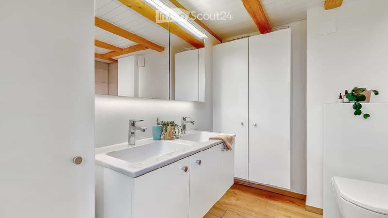 Modern bathroom with white cabinets, double sink, mirror, and a toilet. Wooden floor and exposed wooden beams on the ceiling.