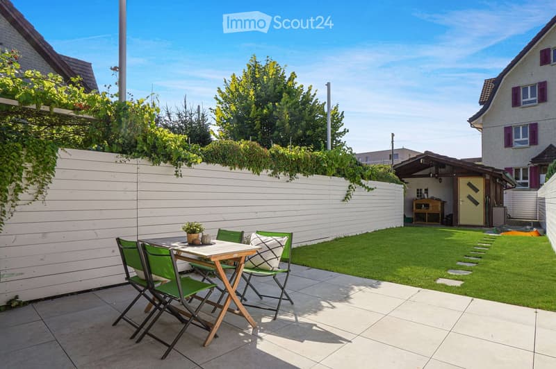 A well-maintained garden with a tiled patio area, a small table with four green chairs, and a potted plant. The garden features a neatly trimmed lawn, a white wooden fence, and a small shed in the background.