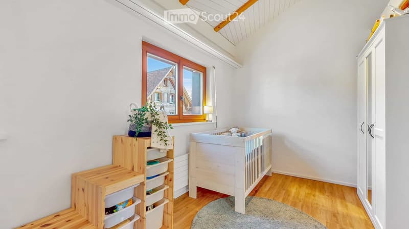 Bright, airy bedroom with wooden flooring, large window, crib, storage unit, plant, and rug.