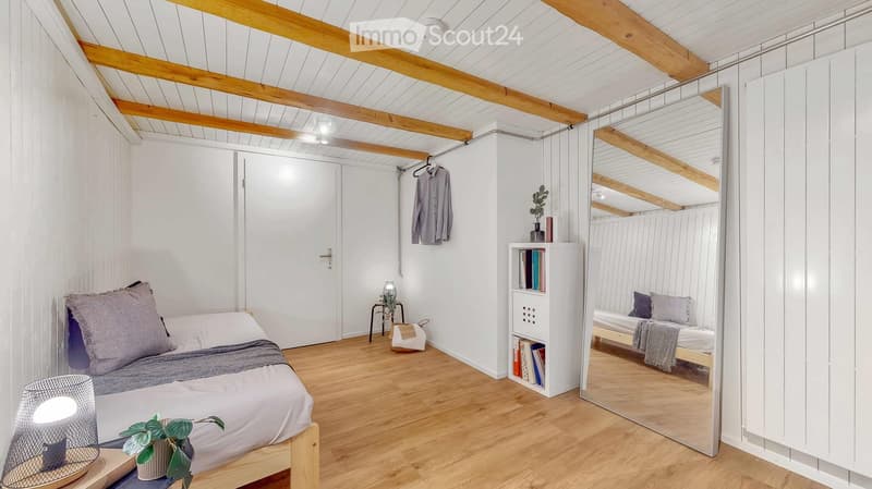 Modern, minimalist bedroom with wooden floors, exposed wooden beams, a bed with grey bedding, a nightstand with a lamp, a large mirror, a bookshelf, and a small plant.