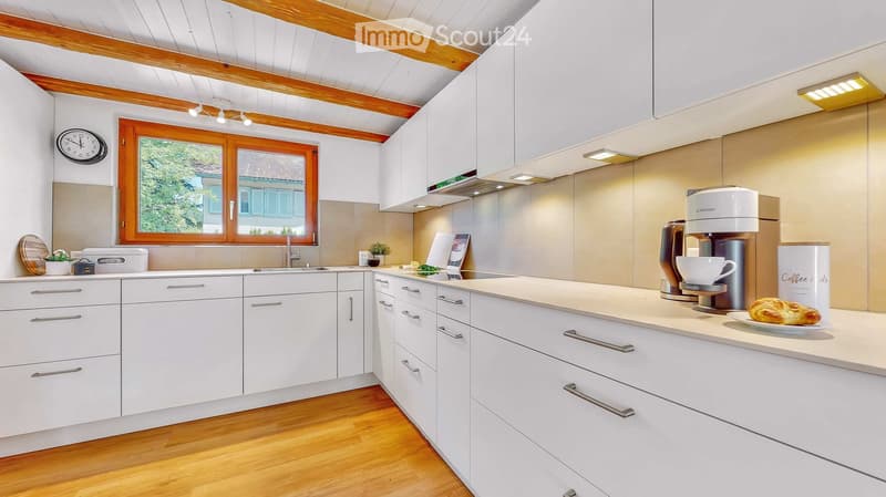 Modern kitchen with white cabinets, wooden ceiling beams, and stainless steel appliances. Features a coffee maker, pastry, and a potted plant on the counter.