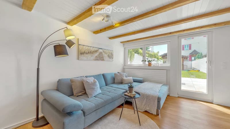 Bright living room with light blue sectional sofa, wooden floor, large windows, and a floor lamp. A small coffee table holds books and a potted plant, with a cozy throw on the sofa.