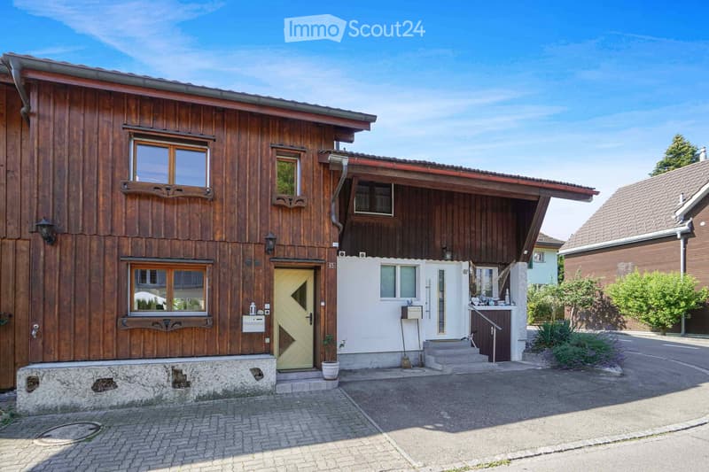 Two-story wooden house with a white entrance section, multiple windows, and a small garden area.