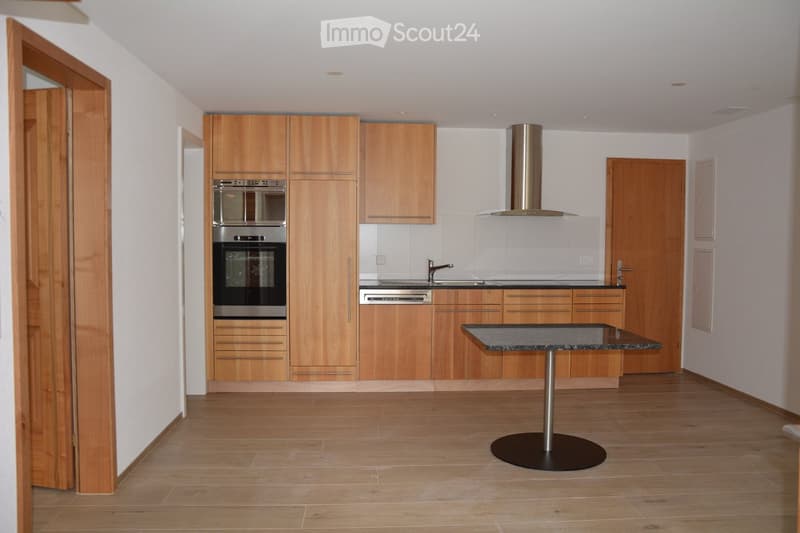 Modern kitchen with light wooden cabinets, stainless steel appliances, a central island with a black countertop, and a round table with a single pedestal.