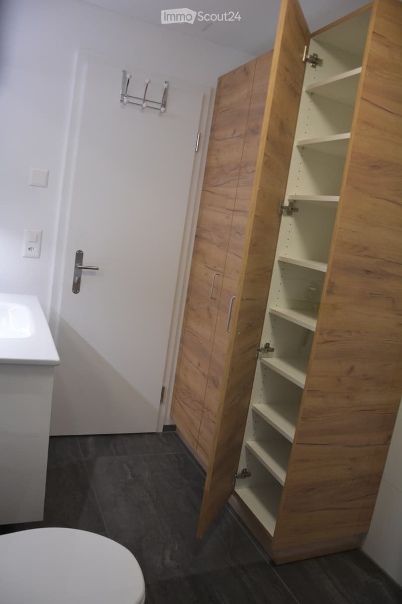 Bathroom with dark tiled floor, white toilet, wooden cabinet with open shelves, coat rack, and door.