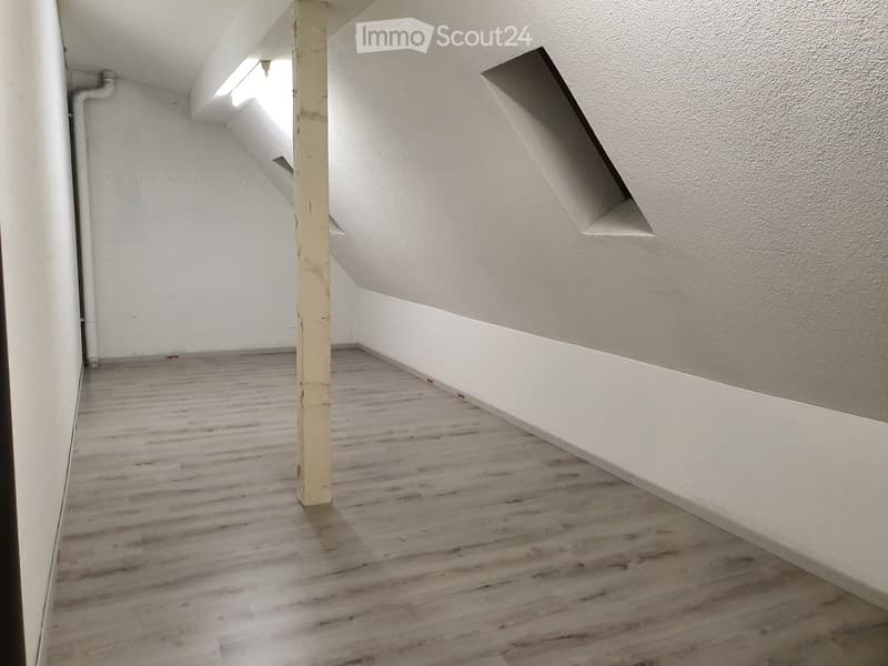 Empty room with wooden flooring, white walls, and a central support column. The ceiling has exposed beams and a skylight window allowing natural light.