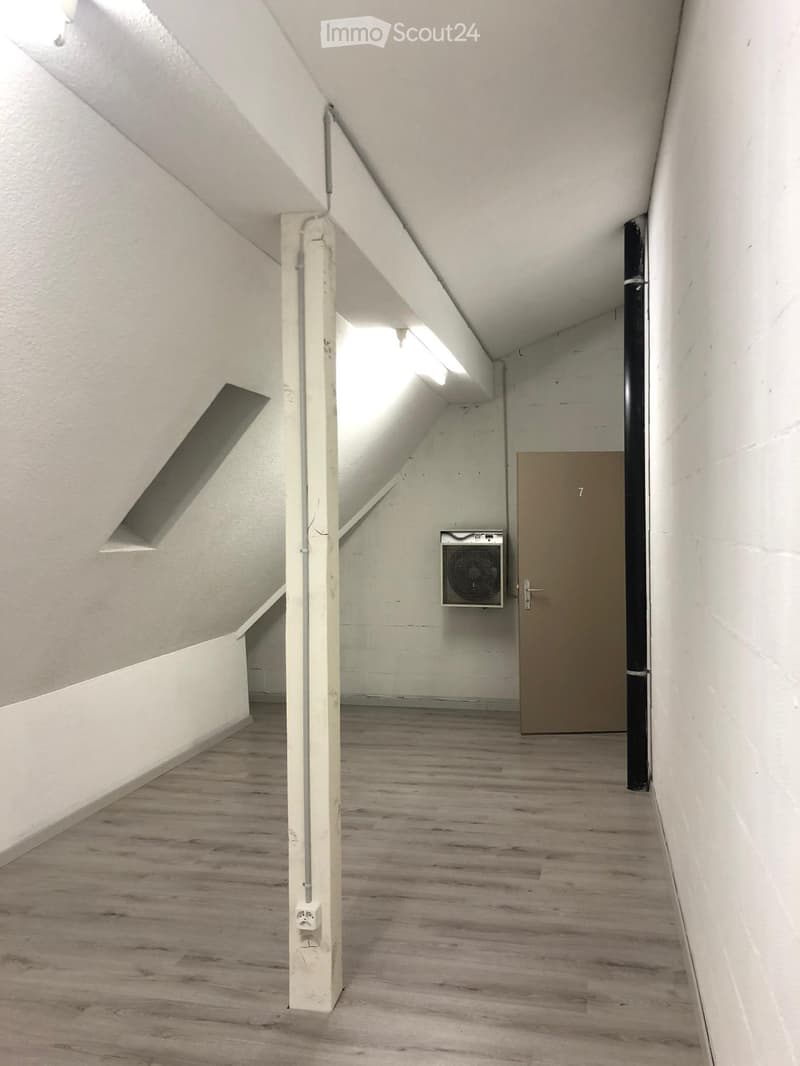 Empty attic space with wooden flooring, white walls, and a single door leading to another room. Features exposed wooden beams and a small ventilation unit on the wall.