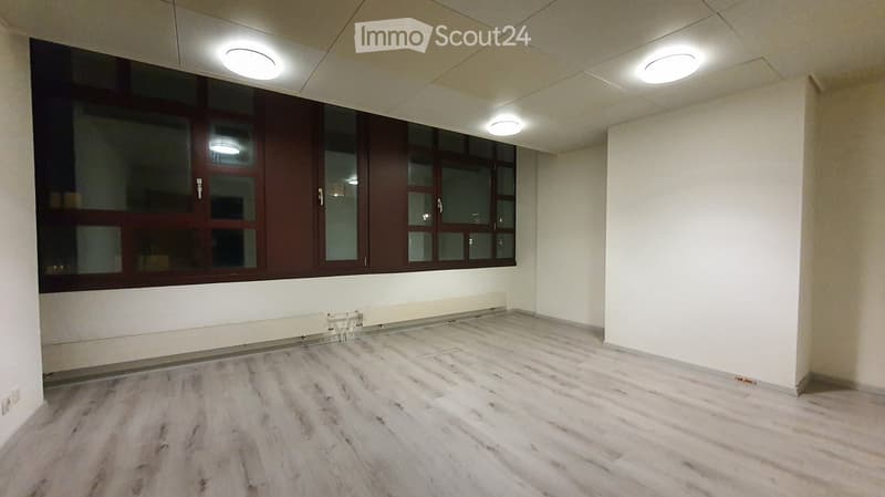 Empty room with light wooden flooring, white walls, large window with dark frame, and recessed ceiling lights.