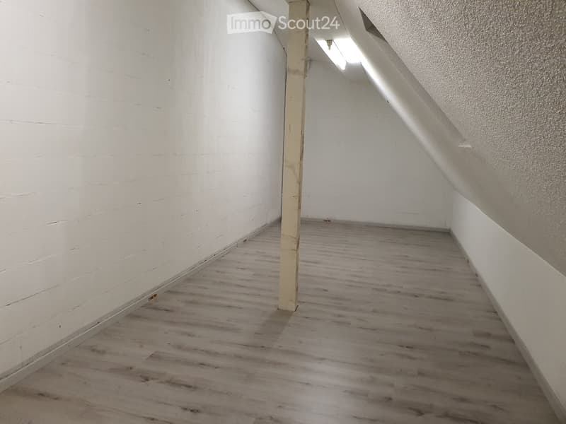 Empty room with wooden flooring and white walls, featuring a single column and a slanted ceiling with exposed beams.