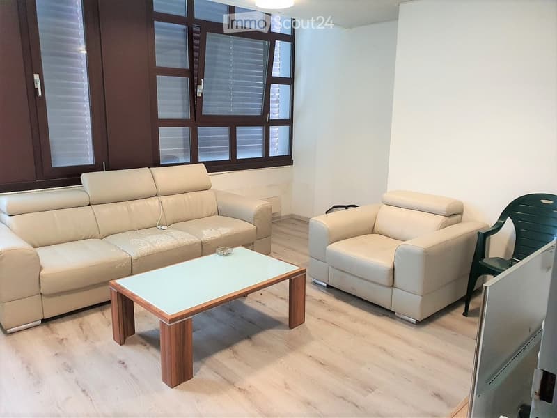 Modern living room with light-colored furniture, including a sofa, armchair, and a glass-top coffee table, set against a backdrop of large windows with blinds.