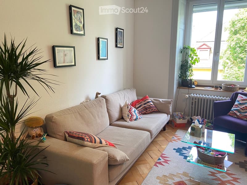 Living room with a beige sectional sofa, colorful throw pillows, a glass coffee table, a purple armchair, a large plant, and several picture frames on the wall.