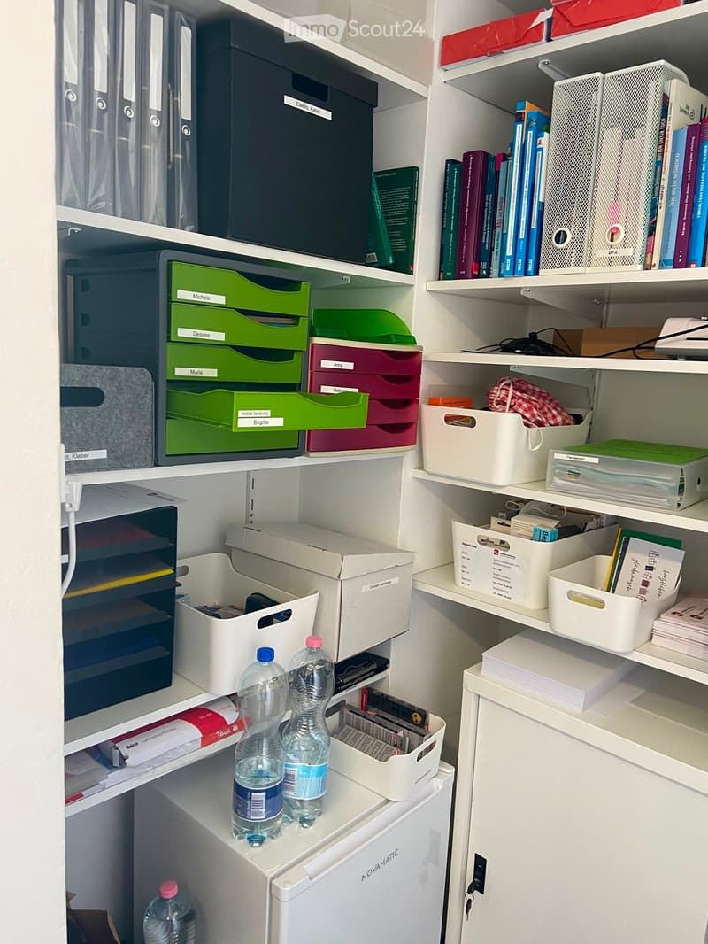 Organized storage area with white shelves, various storage bins, books, and water bottles on a small refrigerator.