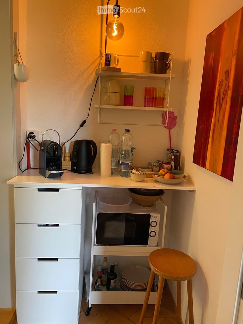 Compact kitchen area with a white countertop, coffee maker, kettle, microwave, and various kitchen items including cups, bowls, and bottles. A wooden stool is placed next to the counter.