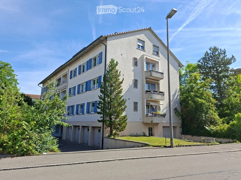 A multi-story residential building with white walls, blue shutters, and balconies. The building features a raised ground floor and is surrounded by greenery and trees.