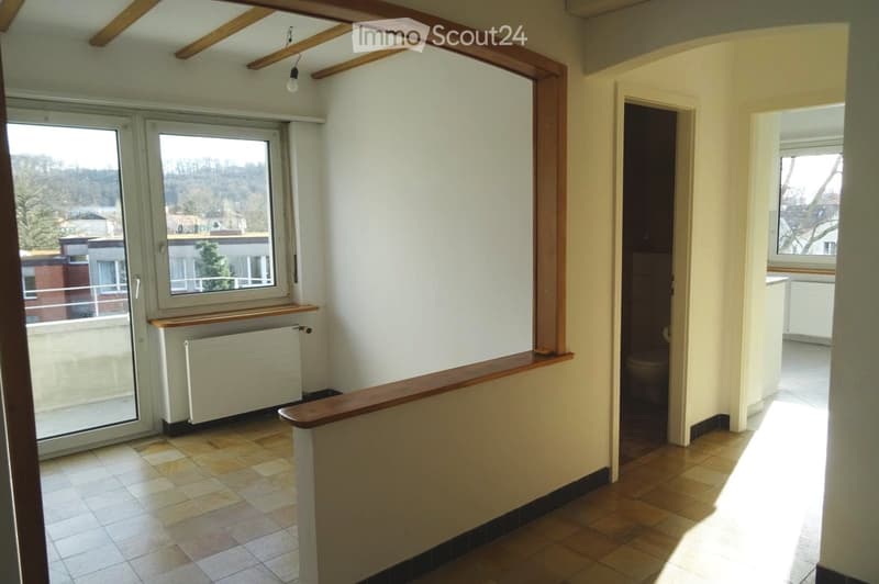 Empty hallway with tiled flooring, wooden railing, and a radiator. The hallway leads to a bathroom with a toilet visible in the background. Natural light streams in from a window on the left side.