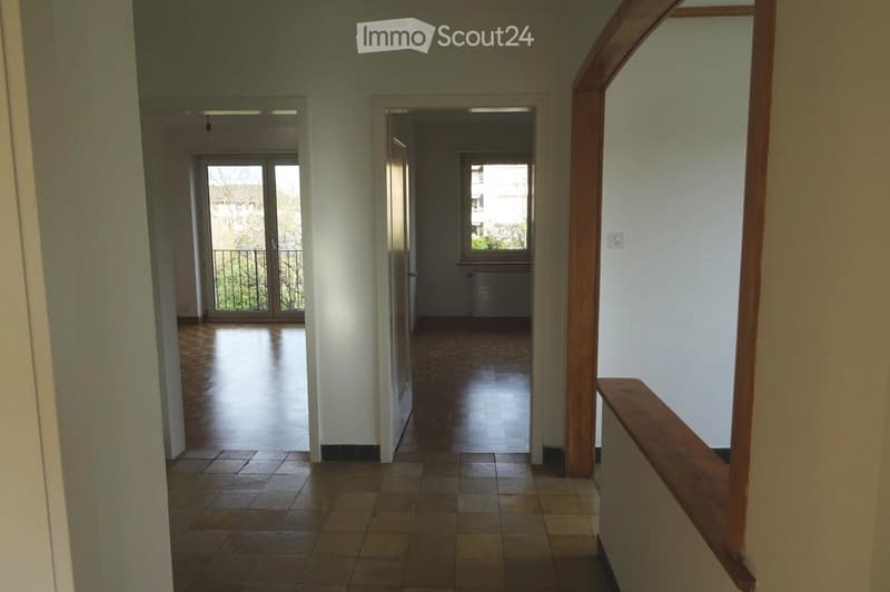 Empty hallway with tiled floor, wooden railing, and doors leading to rooms and a balcony area.