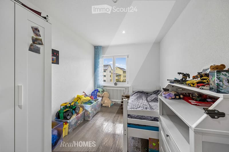 A child's bedroom with a bed, toys, and storage boxes on the floor. A white wardrobe is on the left side, and a white shelf is on the right side. A radiator is fixed to the wall, and a window with a blue curtain is on the wall.