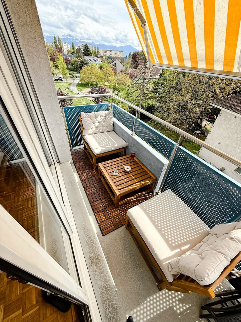 Balcony with wooden decking, sofa, table, and chair, overlooking a scenic view of mountains and greenery.