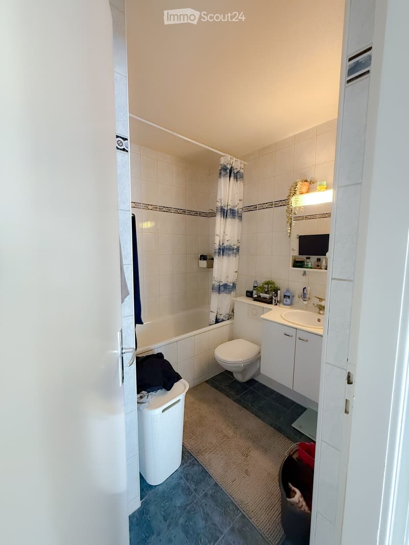 Bathroom with a bathtub, toilet, sink, and cabinet. Features a shower curtain, mirror, and various toiletries on the sink counter.