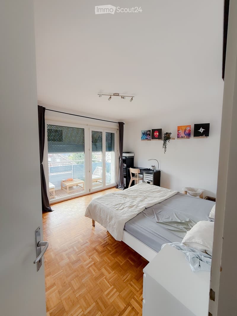 Modern bedroom with wooden flooring, a double bed, a desk, a chair, and a balcony accessible through large glass doors.