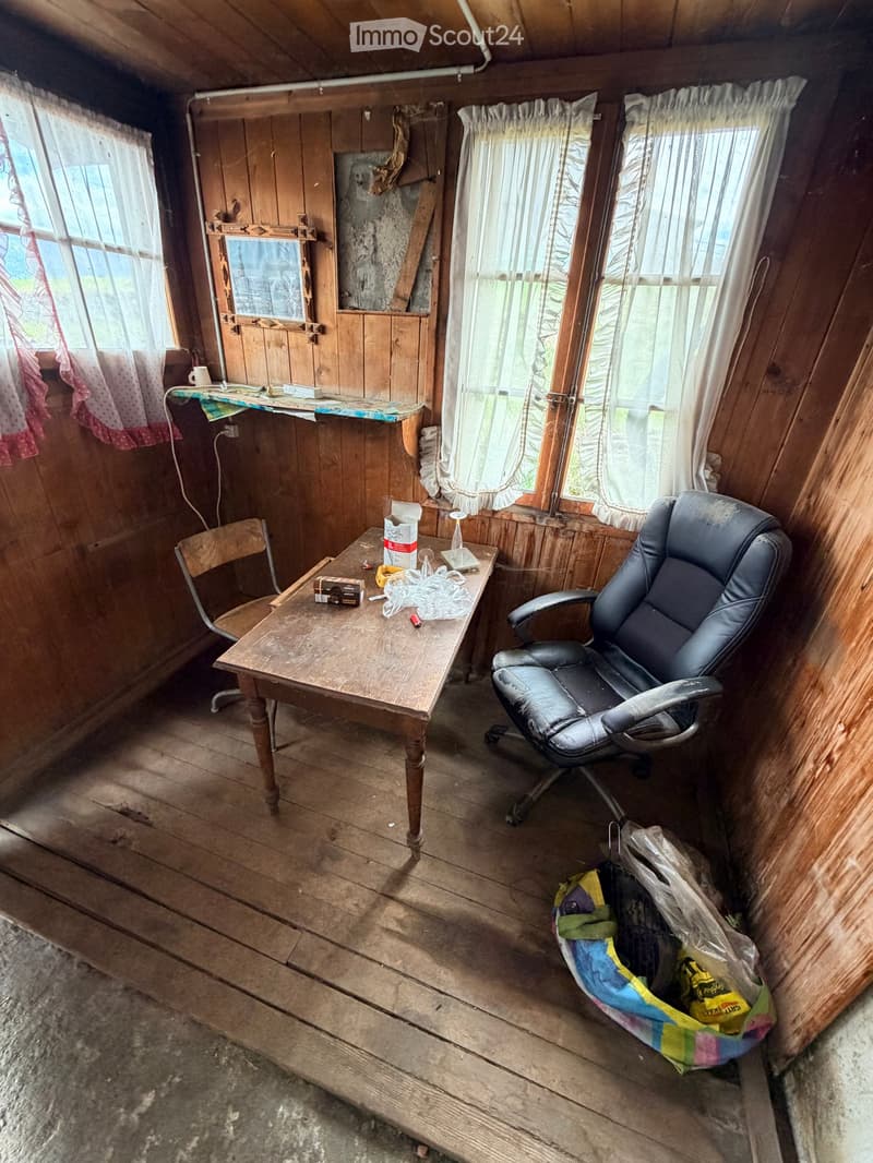 A small, rustic room with wooden walls and floors, featuring a wooden table, office chair, and a plastic bag on the floor.