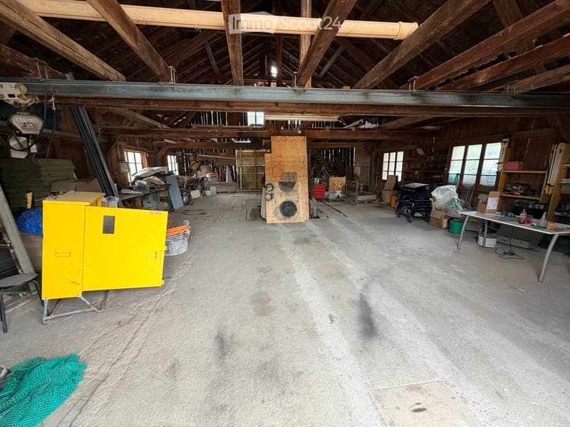 Large, open, unfinished space with exposed wooden beams and concrete floor, featuring various tools, equipment, and storage items scattered throughout.