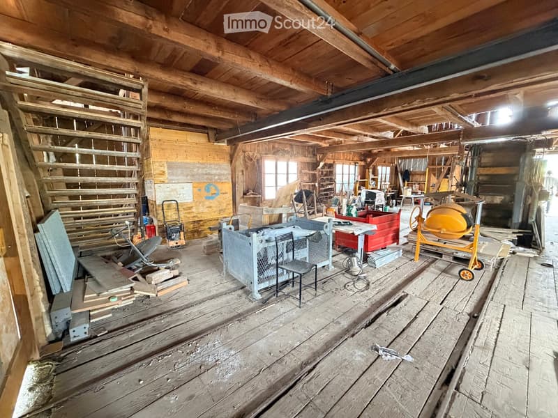 An empty, unfinished room with exposed wooden beams and flooring, various construction materials and tools scattered around, and large windows allowing natural light to enter.