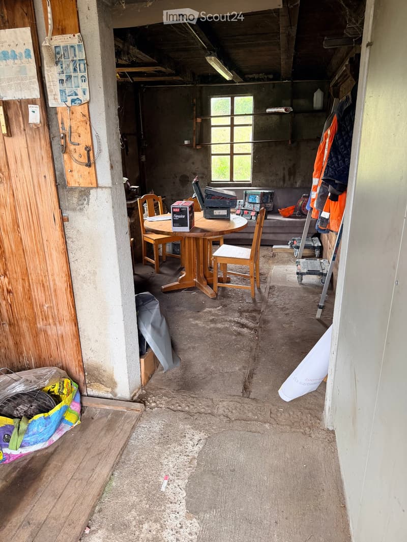 A partially furnished room with a round wooden table, chairs, a sofa, and various storage boxes. The floor is unfinished concrete, and the walls are unfinished concrete with exposed beams. There is a window with bars and a door with a wooden panel on the