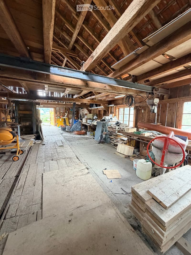 An empty, unfinished attic space with exposed wooden beams and rafters, concrete floor, and scattered construction materials and tools.