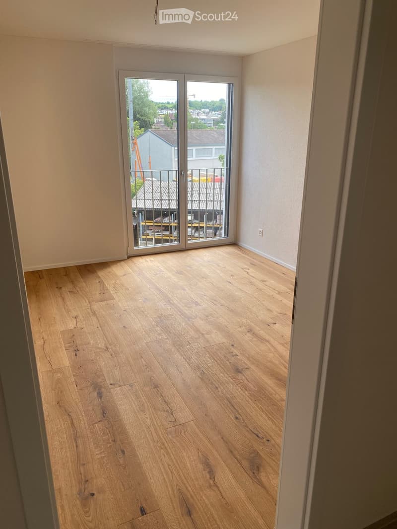 Empty room with light wooden flooring and white walls, featuring a large sliding glass door leading to a balcony with outdoor furniture visible.