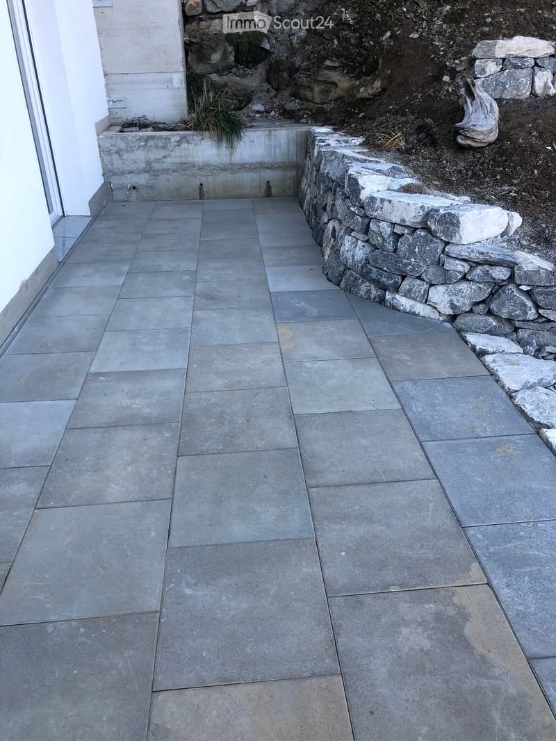 Concrete tiled pathway bordered by a stone wall, with a small concrete planter and some greenery visible on the left side.