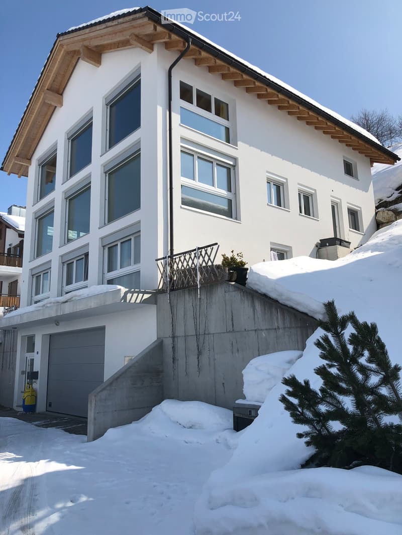 Modern two-story house with white exterior, large windows, wooden roof beams, and a garage. Snow covers the ground and part of the roof.