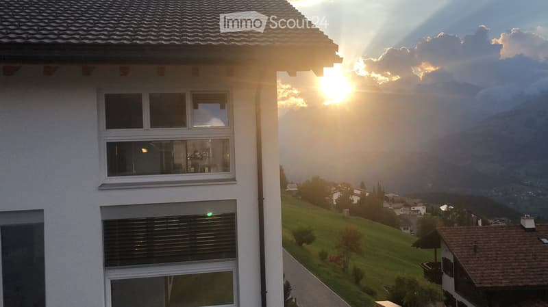 A modern white building with large windows, overlooking a scenic valley with mountains and a setting sun.