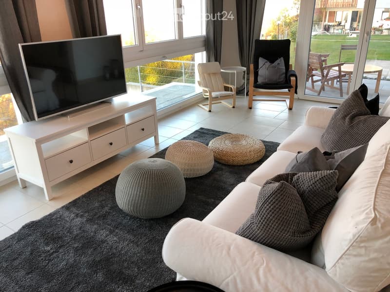 Modern living room with a white sofa, grey throw pillows, a white TV stand, and a large flat-screen TV. The room features a dark grey rug, several round stools, and a black armchair. Large windows and sliding glass doors lead to a balcony with outdoor fur