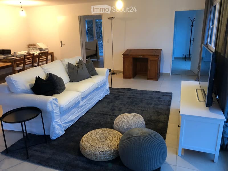 Modern living room with a white sofa, decorative pillows, a dark gray rug, three round woven stools, a small black side table, a white TV stand, and a dining area with chairs and a table visible in the background.