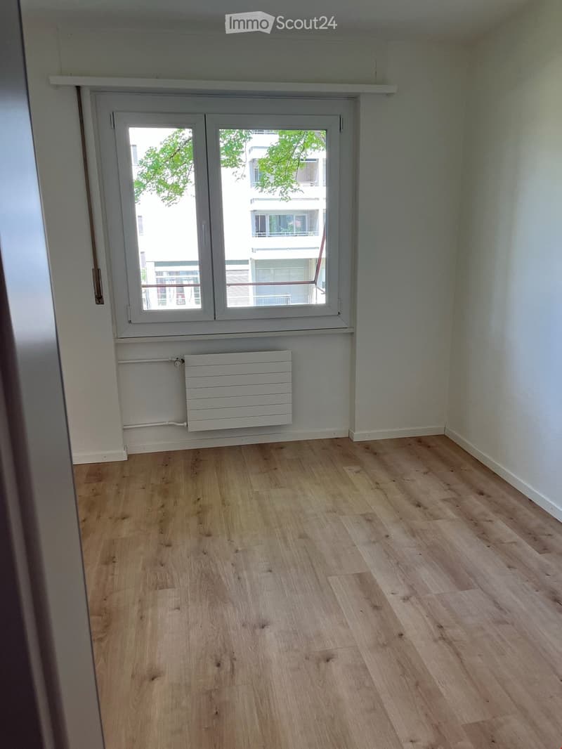 Empty room with light wooden flooring, white walls, and a large window with a radiator below it.