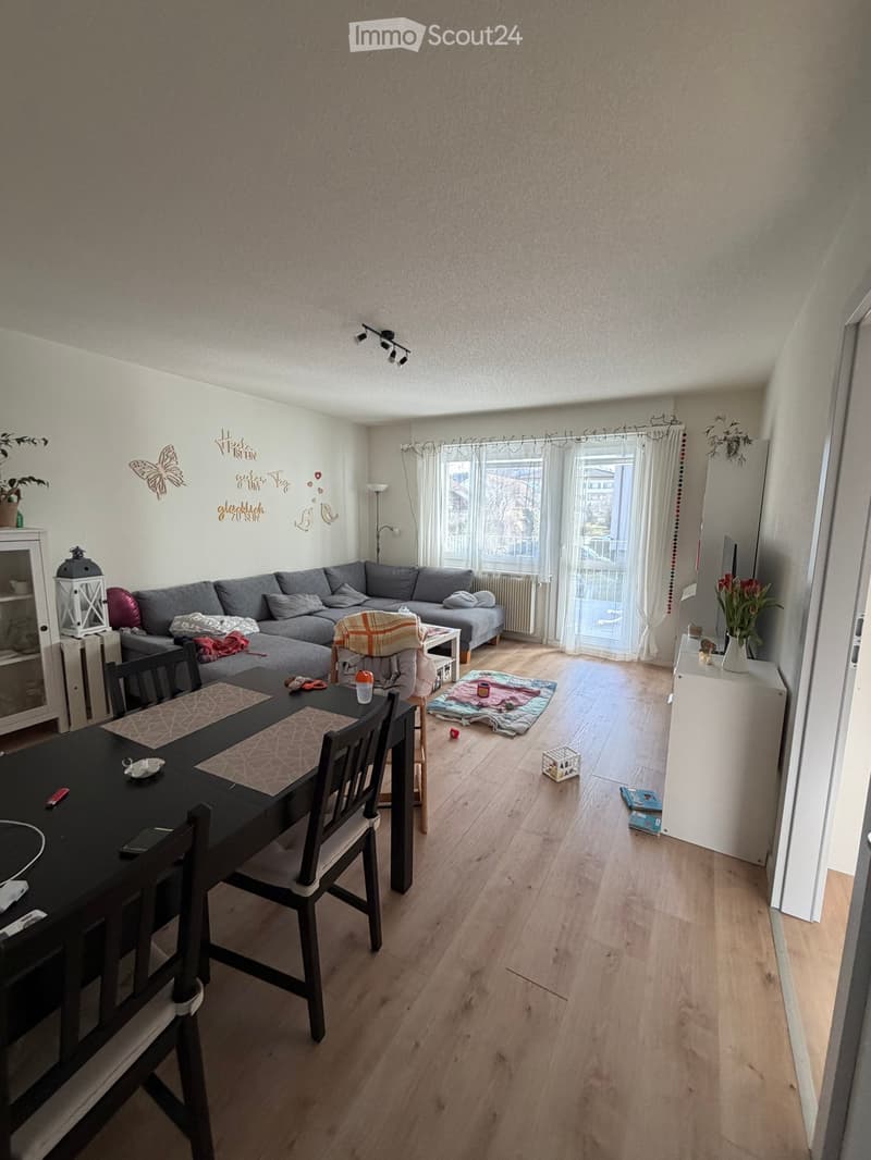 A modern living room with a grey sectional sofa, a black dining table with chairs, a coffee table, a small shelf, and a lamp. The room features wooden flooring, large windows with curtains, and decorative wall art.