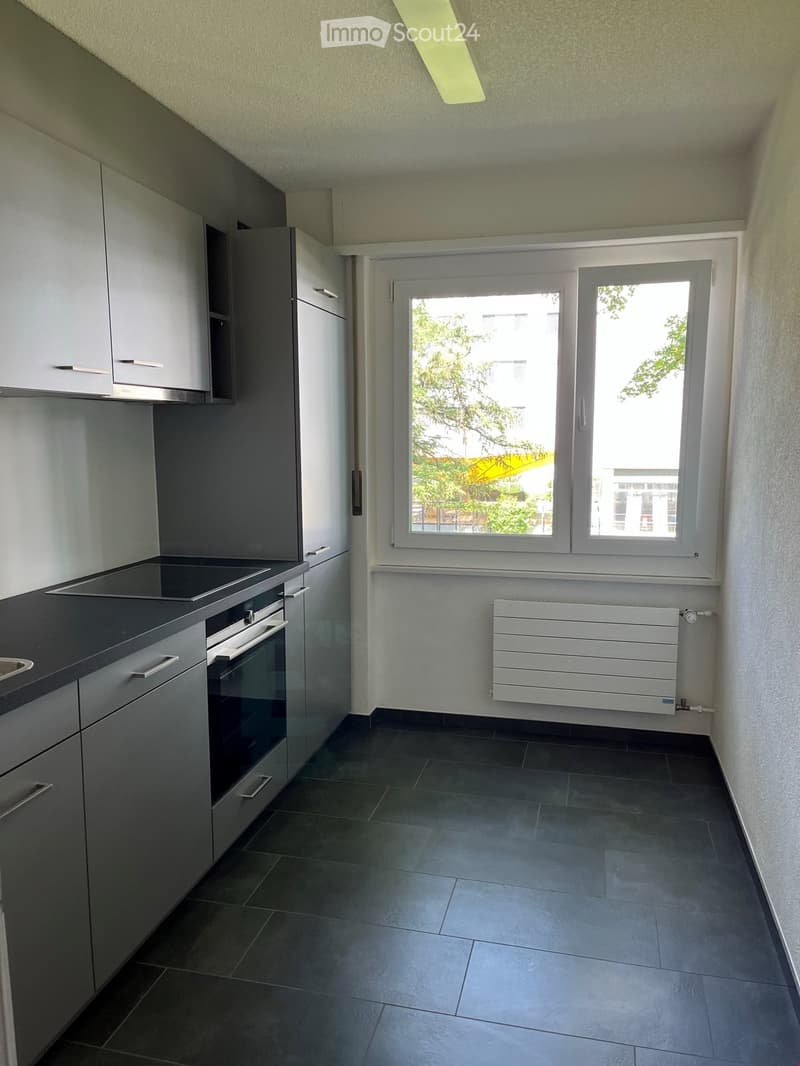 Modern kitchen with grey cabinets, a built-in oven, stove, and radiator. Dark tiled floor and large windows provide natural light.