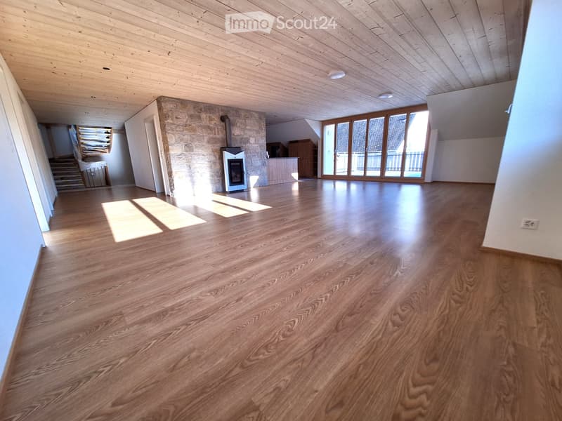 Empty room with wooden flooring, exposed wooden ceiling, and large windows with wooden frames. A stone wall with a fireplace is visible, along with a staircase leading to another level.