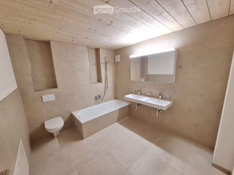 Modern bathroom with beige tiles, a bathtub, toilet, double sink, and a large mirror with built-in lighting.
