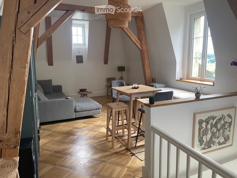 Open-plan living and dining area with wooden flooring, exposed wooden beams, a grey sofa, a coffee table, a dining table with chairs, and a small bed area with a window seat.